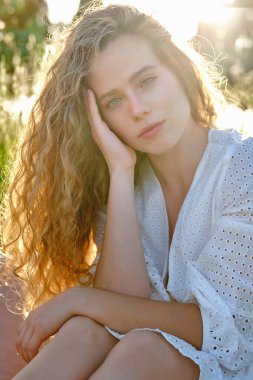 Arkadaki genç kadın beyaz bir elbise giyiyor yanaklarına dokunuyor ve yeşil güneşli doğada otururken kameraya bakıyor.