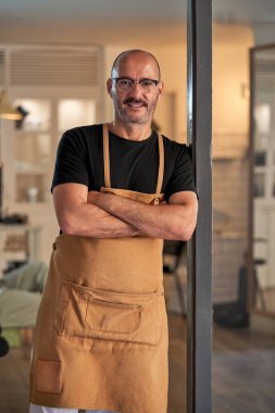 Önlüklü, gözlüklü, çapraz elli ve modern berber dükkanında çalışırken kameraya bakan kendinden emin bir erkek.