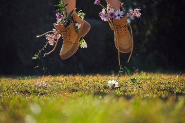 Tanımlanamayan kişi kahverengi çizmeler ve kır çiçekleri giyerek güneşli yaz gününde yeşil bitkilere karşı doğada çimlere atlıyor.