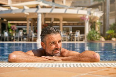 Neşeli sakallı orta yaşlı erkek turist yaz tatili sırasında temiz mavi suda dinlenirken yüzme havuzunun kenarına yaslanıyor.