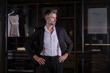 Yetişkin, orta yaşlı, şık elbiseli bir erkek gardıroba karşı duruyor ve elleri belinde başka yere bakıyor.