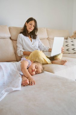 Gencecik bir anne dizüstü bilgisayarı karıştırıyor ve evdeki oturma odasında uyuyan küçük bir kızın yanında otururken uzaktan çalışıyor.
