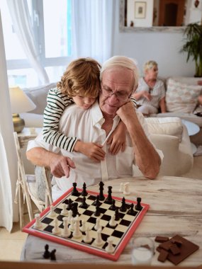 Küçük çocuk yaşlı adama sarılıyor ve büyükannesi evdeki rahat oturma odasında kanepede otururken masada satranç oynamayı öğreniyor.
