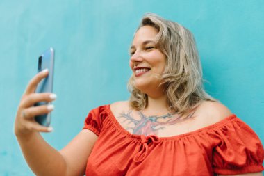 Mavi duvar arka planında dikilirken akıllı telefonuyla selfie çeken gündelik kıyafetli genç ve iri kadın portresi.