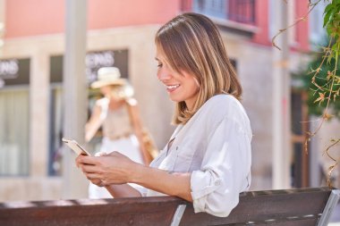 Beyaz bluzlu, ahşap bankta dinlenen ve güneş ışığıyla aydınlatılan şehir caddesinde cep telefonu kullanan gülümseyen kadının yan görüntüsü