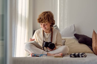 Profesyonel fotoğraf makinesiyle gülümseyen okul çocuğu evde bacak bacak bacak üstüne atmış otururken not defterine not alıyor.