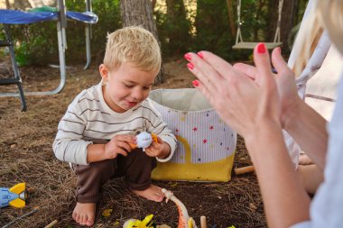 Bluzlu bir kadın, yeşil ağaçlarla parktaki popoların üzerinde otururken oyuncaklarla oynayan sevimli çocuğu alkışlıyor.