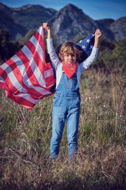 Çimenli çayırlarda elinde Amerikan bayrağı sallayan ve kameraya bakan kot pantolonlu, genç ve tatlı bir çocuk.