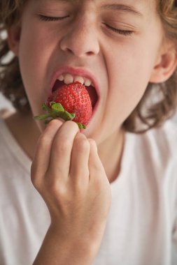 Beyaz tişörtlü mutlu çocuk tatlı vitamin meyvesi yerken sevinçle gözlerini kapıyor.