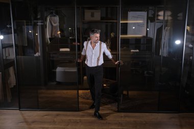 Beyaz gömlekli, siyah pantolonlu, orta yaşlı sakallı bir adam soyunma odasının kapısında duruyor.