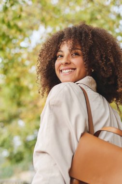 Kıvırcık saçlı neşeli Afro-Amerikan kadın ağaçların bulanık arka planında dikilirken gülümsüyor ve başka tarafa bakıyordu.