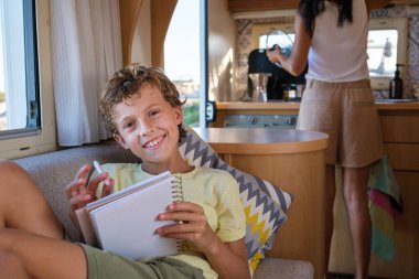 Glad boy with pen and notepad smiling and looking at camera while sitting on sofa near crop mother and making notes in diary during road trip clipart