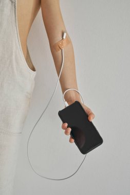 Koluna elektrik kablosu bağlanmış, dijital bağımlı çocuk gri arka planda akıllı telefon olduğunu gösteriyor.