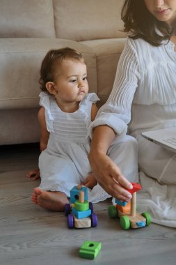 Beyaz elbiseli sevimli küçük kıza oyuncaklarını gösteren tanınmamış bir anne. Oturma odasındaki ahşap zeminde birlikte otururken.