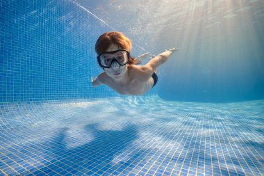 Gözlüklü ve şortlu bir çocuk, gün ışığının üzerinde şeffaf bir suda yüzüyor ve kameraya bakıyor.