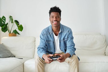 Keyifli, genç, Afro-Amerikan erkek oyuncu rahat bir kanepede joystick ile otururken video oyunu oynuyor.