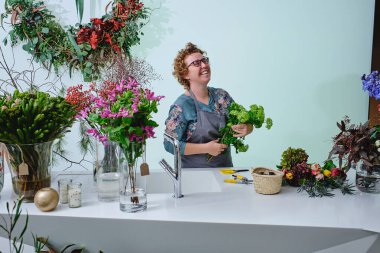 Elinde cam vazolarda çiçek açan tezgahın yanında dikilirken yeşil trakyum buketi düzenleyen pozitif yetişkin kadın çiçekçi.