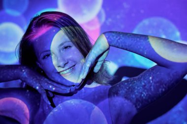 Olumlu genç bir kadın çenesine dokunuyor ve renkli ışık yansımalarının altında dururken kameraya gülümsüyor.