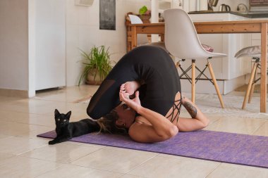 Spor kıyafetli genç bir kadının evde kedinin yanında yoga minderinde poz verişi.