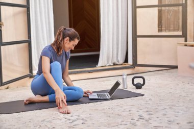 Spor kıyafetleri içinde çıplak ayaklı genç bir kadının bacak bacak üstüne atmış yoga minderinde oturup terasta dizüstü bilgisayarda video izlemesi.