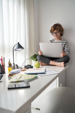 Gündüz vakti internet üzerinden ders alırken masada oturup ekrana bakan çıplak ayaklı zeki bir çocuk.