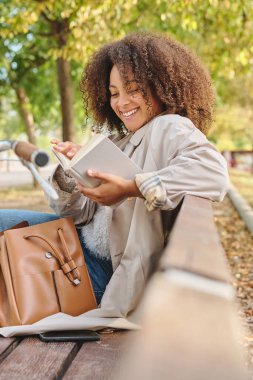 Afro saç stili, sırıtan Afro-Amerikan kız öğrenci tahta bankta çanta ve kitap okuyarak oturuma hazırlanıyor.