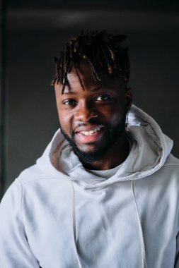 Kendine güvenen, kapüşonlu Afro-Amerikan erkek. Kameraya pozitif yüz ifadesiyle bakıyor. Odada karanlık arka planda duruyor.