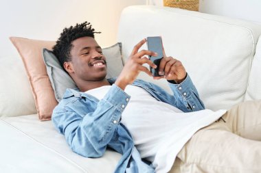 Gülümseyen Afro-Amerikan erkek rahat koltukta uzanıyor ve modern apartmandaki cep telefonlarını karıştırıyor.