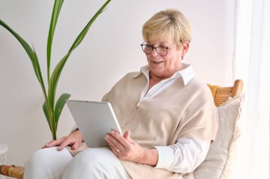 Evde saman sandalyede dinlenirken tablette internette sörf yapan, göz giysisi giyen yaşlı bir kadın.