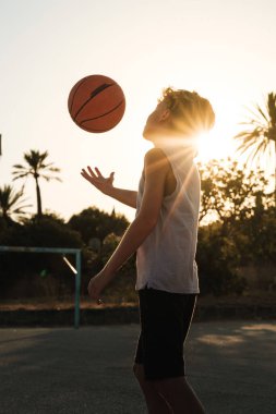Gökyüzüne karşı antrenman yaparken spor sahasında basketbolla oynayan sporcu çocuğun yan görünüşü