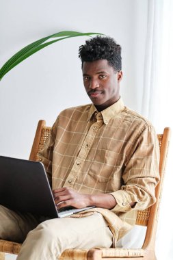 Kapalı alanda saman sandalyede otururken kameraya bakan kareli gömlekli, Afro saçlı genç bir adam.