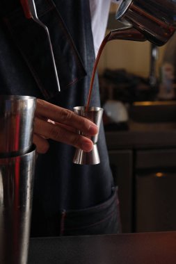 Siyah önlüklü isimsiz bir barmen metal çaydanlıktan taze kahveyi tezgahın yakınına dolduruyor.