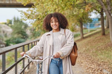 Parkta bisikletle yürürken kameraya bakan kıvırcık saçlı, gülümseyen Afrikalı Amerikalı bir kadının portresi.