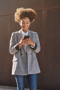 Güneşli bir günde sokakta durup cep telefonuyla mesaj gönderirken Afro saç stili gülümseyen tatmin olmuş zenci kadın.