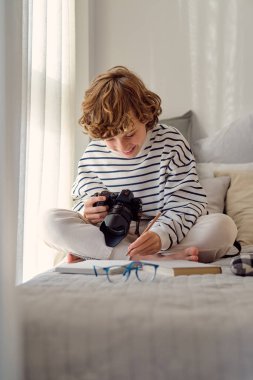 Profesyonel fotoğraf makinesi olan mutlu bir okul çocuğu evdeki not defterinin yanında bacak bacak bacak üstüne atmış bir şekilde oturuyor.