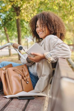 Pozitif Afrika kökenli genç bir Amerikalı kadının park bankında oturması, çantası, kitabı ve başka tarafa bakması.