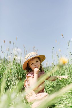 Güneşli bir yaz günü çimenli bir çayırda oturmuş parmağını emerek kameraya bakarken mavi gökyüzüne bakan sevimli küçük bir kız.