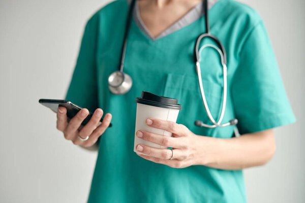 female surgeon in green uniform with stethoscope and takeaway hot beverage browsing internet on cellphone on light background