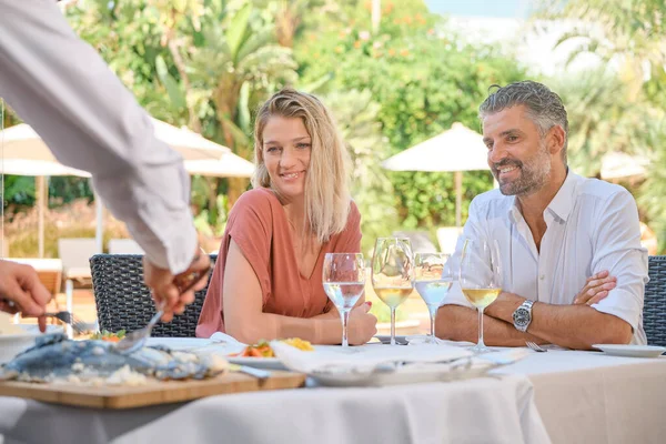 在热带酒店的餐馆度过暑假的时候 微笑的男朋友和女朋友坐在餐桌旁 一边喝着酒杯 一边看着杂烩侍应生端菜 — 图库照片