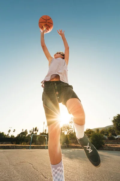 Low angle of boy in activewear throwing orange ball while playing streetball against sunshine on sports ground