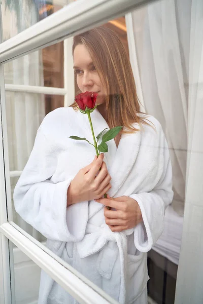 窓に立って思慮深く見つめている白いテリーバスローブの若い夢見る女性の窓を通して — ストック写真
