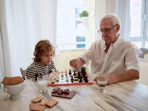 Elderly Man Teaching Concentrated Preteen Boy Play Chess While Sitting — Stock Photo, Image