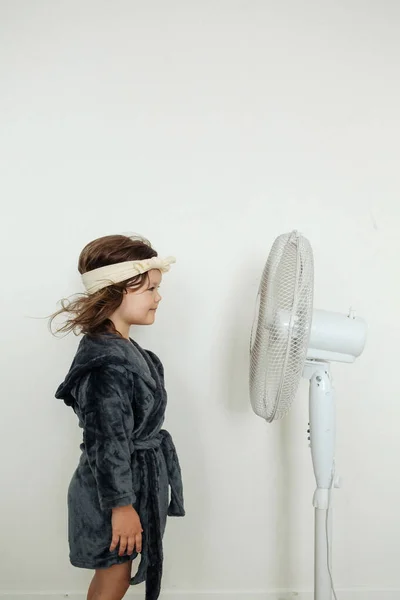 熱い日の間に部屋の白いファンに対して立っている灰色のローブおよびヘッドバンドを身に着けているかわいい少女の側面の眺め — ストック写真