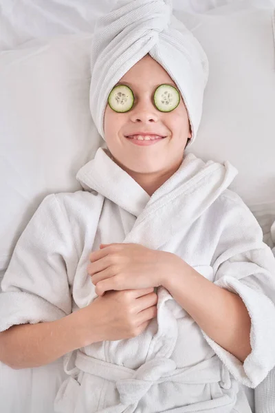 穿着白色浴衣 头戴毛巾 面带微笑的孩子在家里做日常护肤活动 一边在家中用黄瓜片放松一下 一边玩乐 — 图库照片