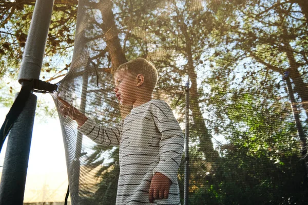 木の晴れた日に遊び場で遊んでいる間トランポリン網に触れる少年の側面の眺め — ストック写真