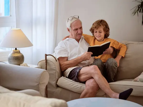 老年人和心满意足的孙子一起看书 在舒适的客厅里舒服的沙发上躺在一起 — 图库照片