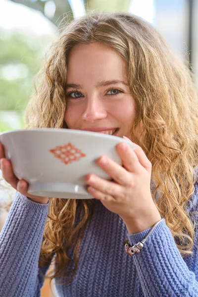 迷人的卷发女人穿着针织毛衣 看着相机 喝着陶瓷碗里的拉面汤 在亚洲餐馆里呆着 — 图库照片