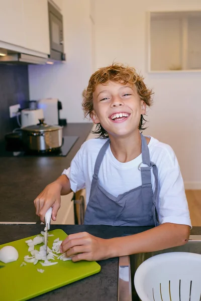 Cheerful child in apron cutting raw onion with knife on chopping board while looking at camera in house kitchen