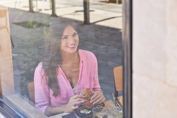 通过对穿着粉色休闲衫 一头长长的深色头发坐在窗边餐桌旁 午餐在咖啡店吃汉堡包的积极女青年的玻璃透视 — 图库照片