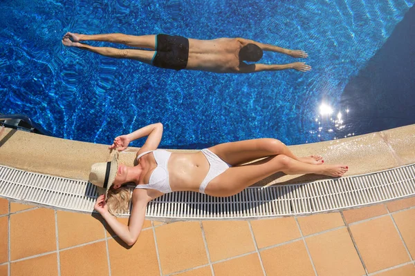 From above of anonymous woman in white bikini and hat sunbathing while man swimming in pool at tropical resort on summer vacation